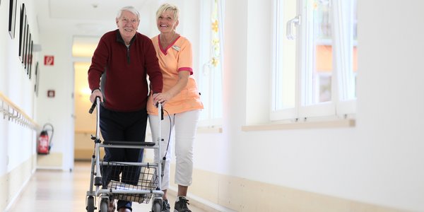 Eine Pflegefachkraft hilft einem Bewohner, der mit dem Rollator im Flur unterwegs ist.